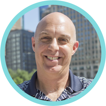 A smiling, bald man outdoors in an urban setting with tall buildings in the background.