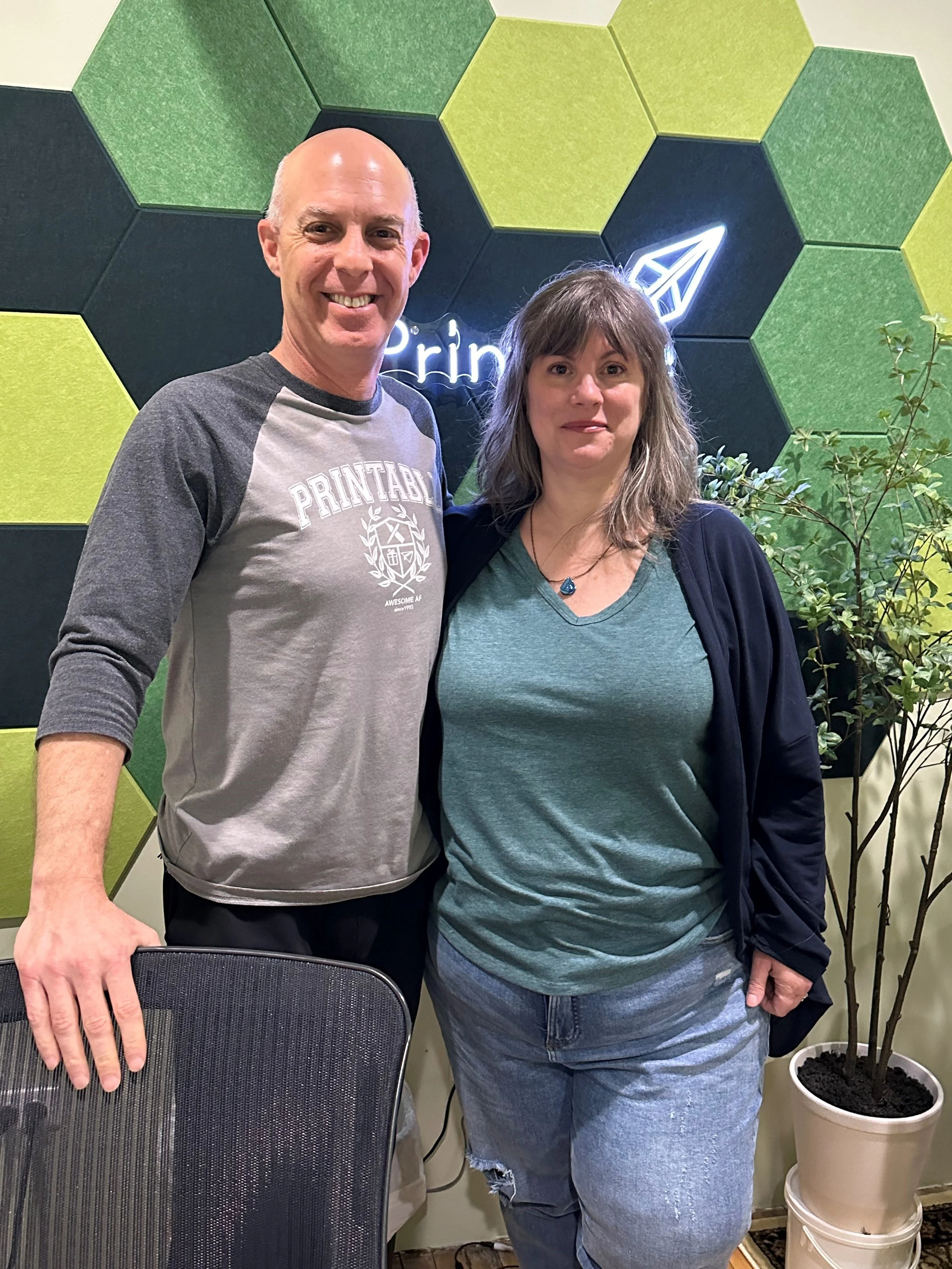 A man and a woman stand side by side indoors, smiling at the camera. The man wears a gray and black long-sleeve shirt with 'PRINTABLES' on it, and the woman wears a teal t-shirt and a black jacket. They are in front of a wall with hexagonal green and