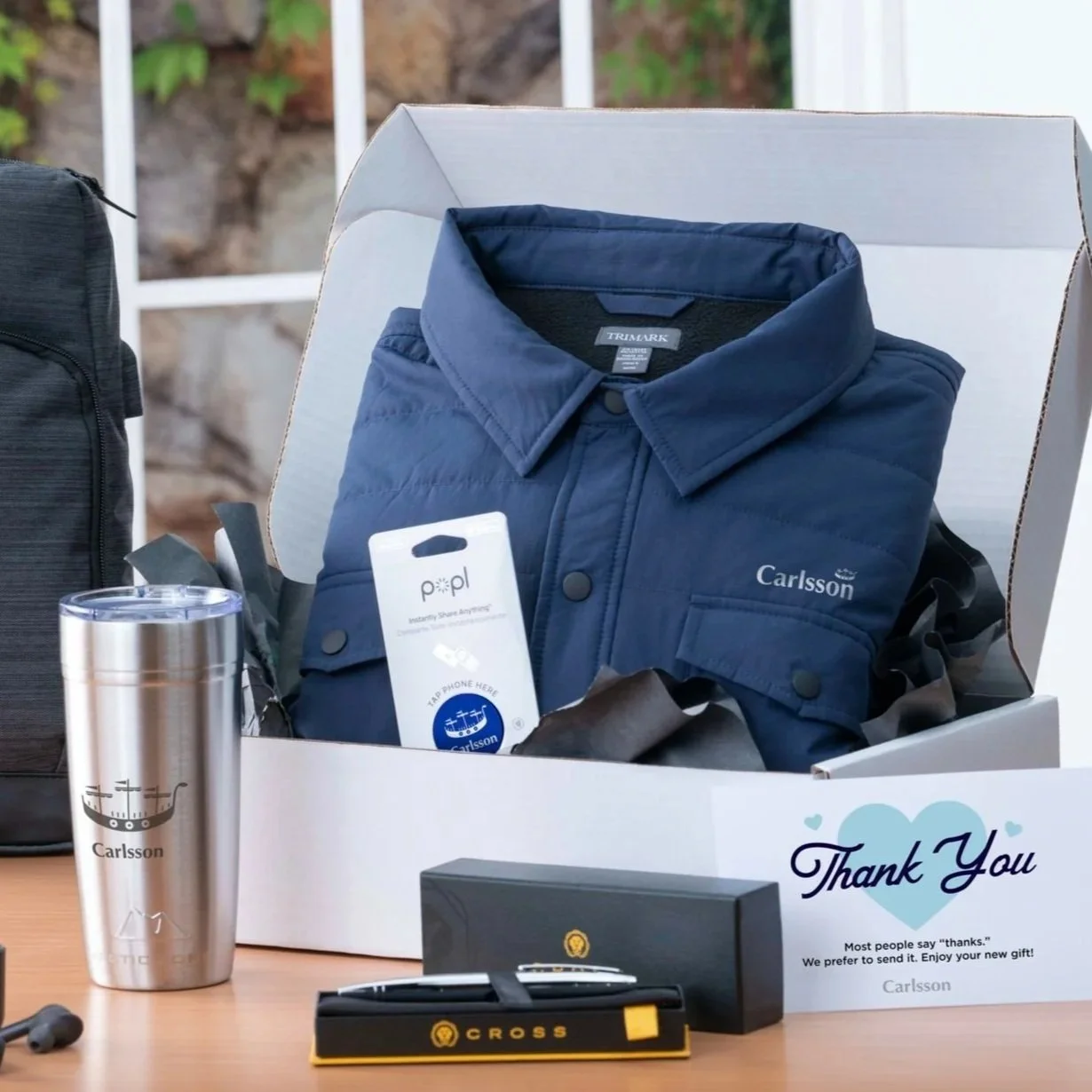 A gift box containing a blue jacket embroidered with the name 'Carlsson,' a scannable tag, and a thank you card. Next to the box is a silver tumbler with the name 'Carlsson' and a ship logo, and a black box and pen labeled 'Cross' on the table.