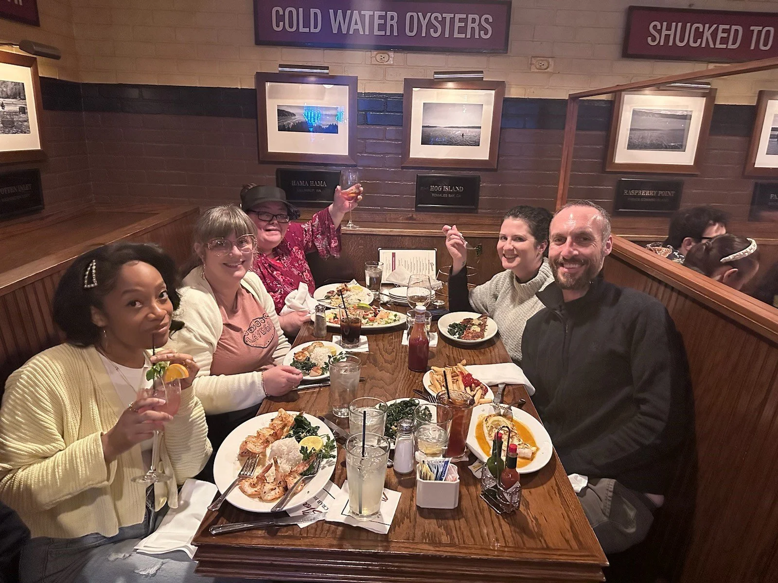 Group of seven people, five women and two men, sitting around a wooden table in a restaurant, enjoying drinks and food, with framed photos and signs on the wall behind them.