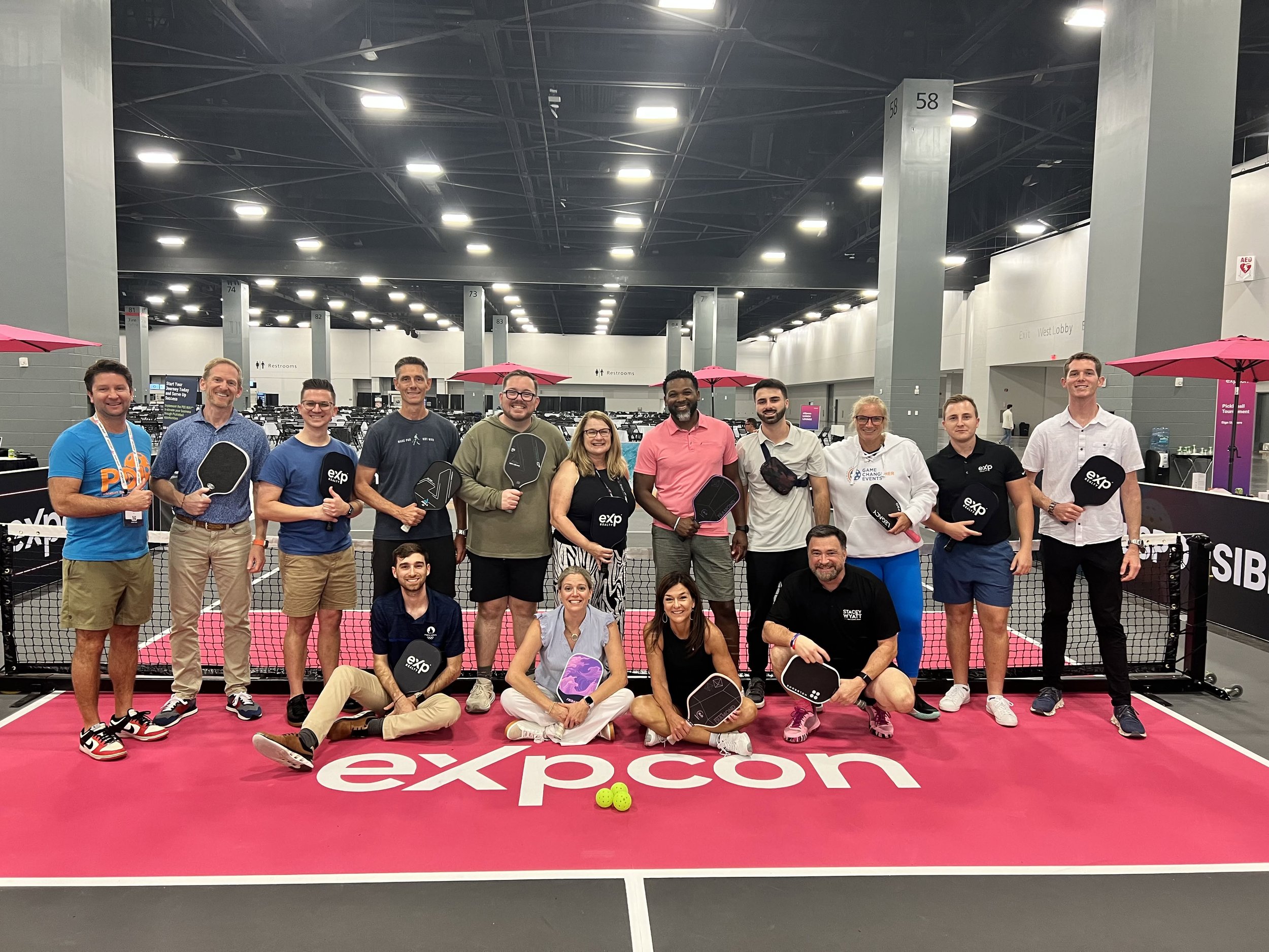 A group of people is standing on an indoor pickleball court with pink flooring. They are holding pickleball paddles, and the word 'expcon' is visible on the floor. The setting appears to be a convention or event space with multiple columns and pink u