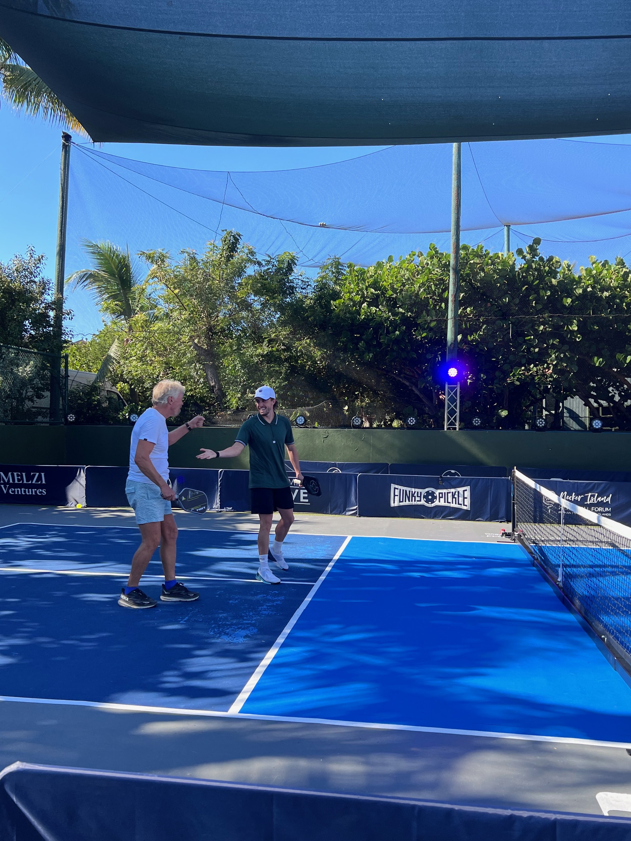 Two people playing pickleball on a blue court, with a shaded canopy overhead and trees in the background.