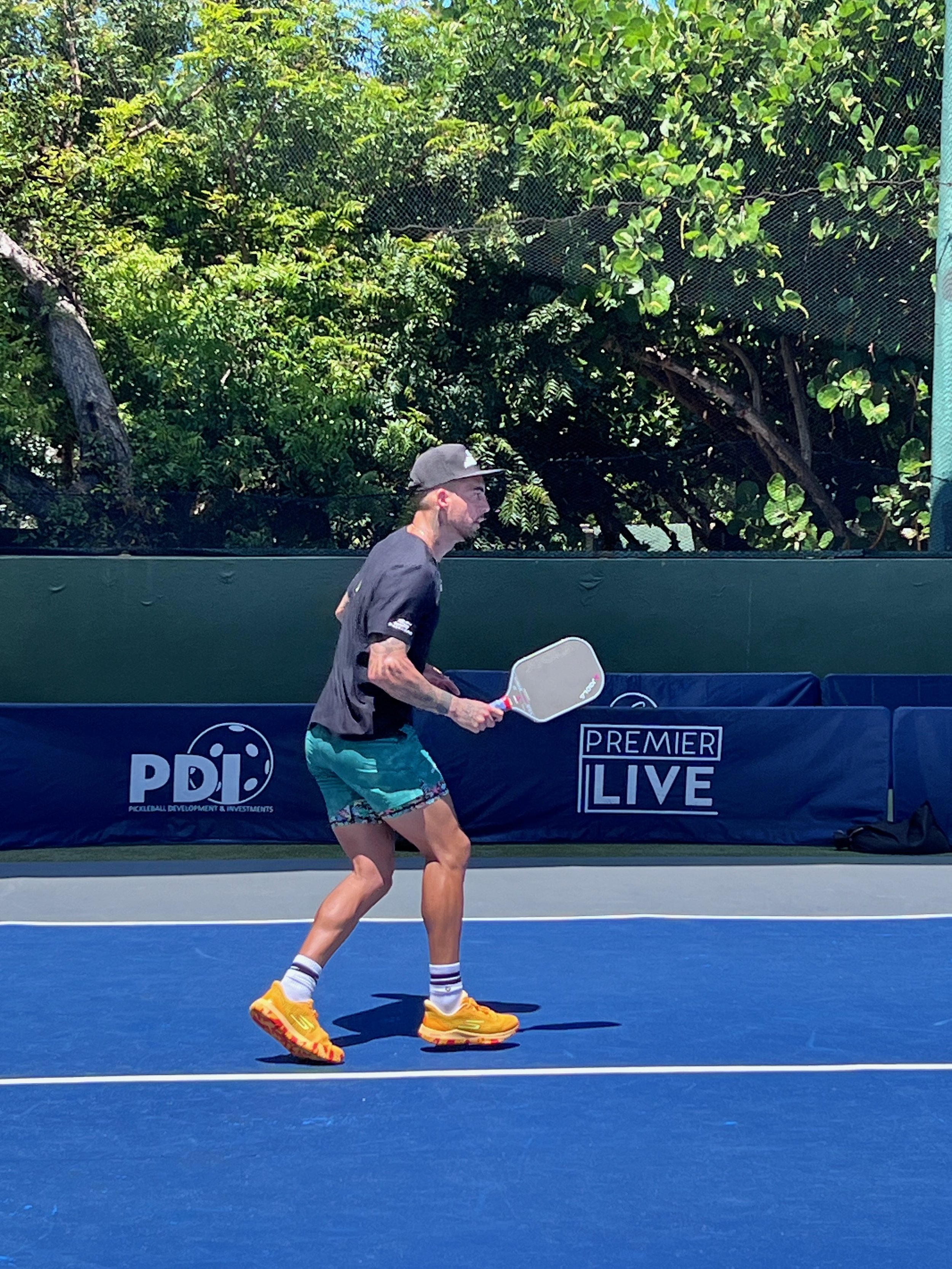 Person playing pickleball on an outdoor court, holding a paddle and wearing athletic gear, including a cap and bright shoes. Background features greenery and a branded banner.