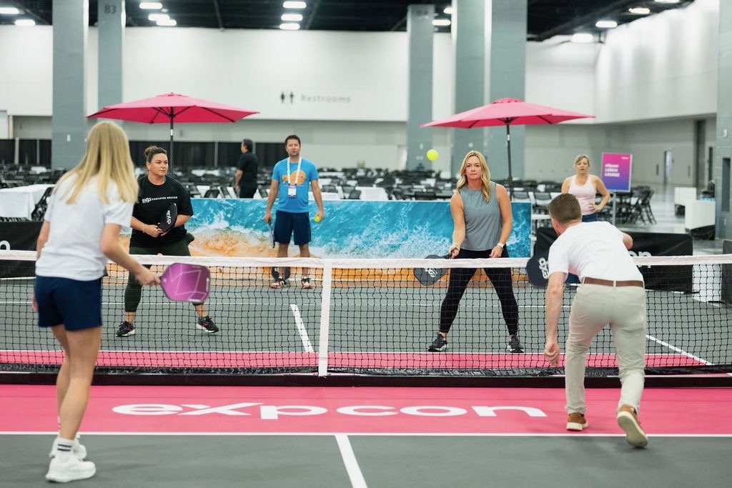 People playing pickleball indoors on a pink court at an expo event, with spectators and banners in the background.