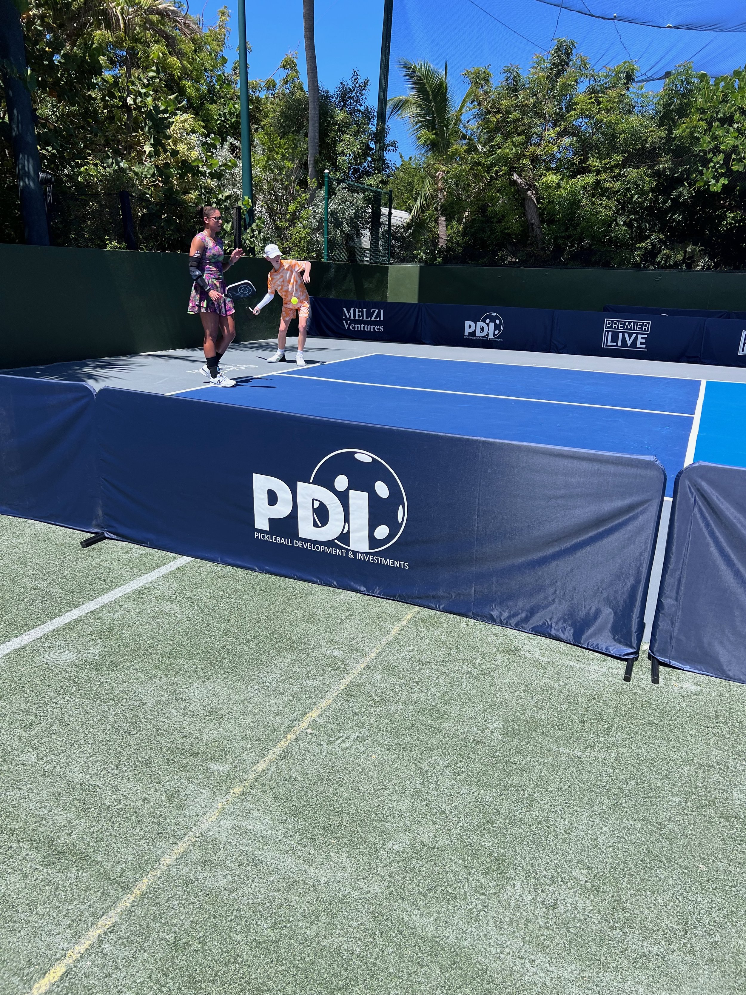 Two people playing pickleball on an outdoor court surrounded by green foliage and blue skies, with a PDI (Pickleball Development & Investments) banner visible.