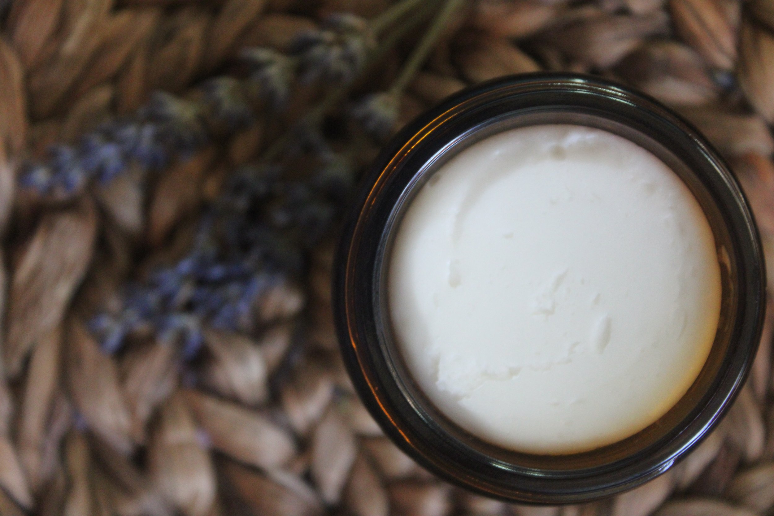 Top view of a glass jar filled with a creamy magnesium lotion, placed on a woven basket with a lavender sprig in the background.