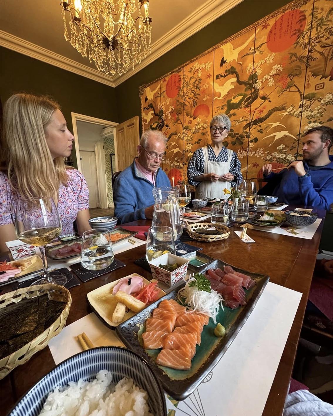 People enjoying a traditional Japanese sushi meal around a wooden dining table in a decorated room with a large ornate chandelier and a colorful wall hanging featuring cranes and stylized trees.