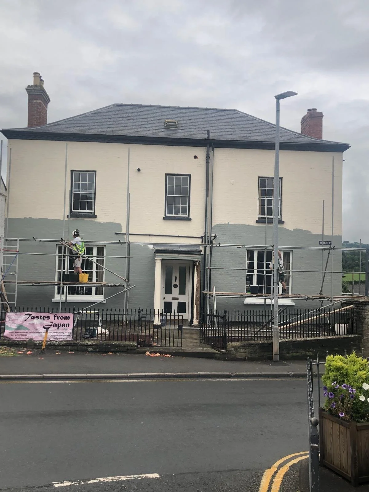 A house undergoing exterior renovation with scaffolding in place. Two workers are painting the house's facade, which is partially painted gray. There is a sign on the fence that reads "Tastes from Japan." The street in front of the house has a flower