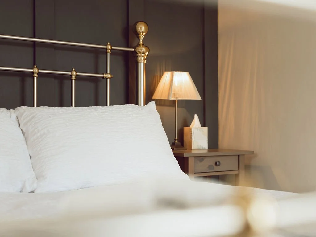 A neatly made bed with white pillows and bedding, a bedside table with a lamp, tissue box, and dark wall background.