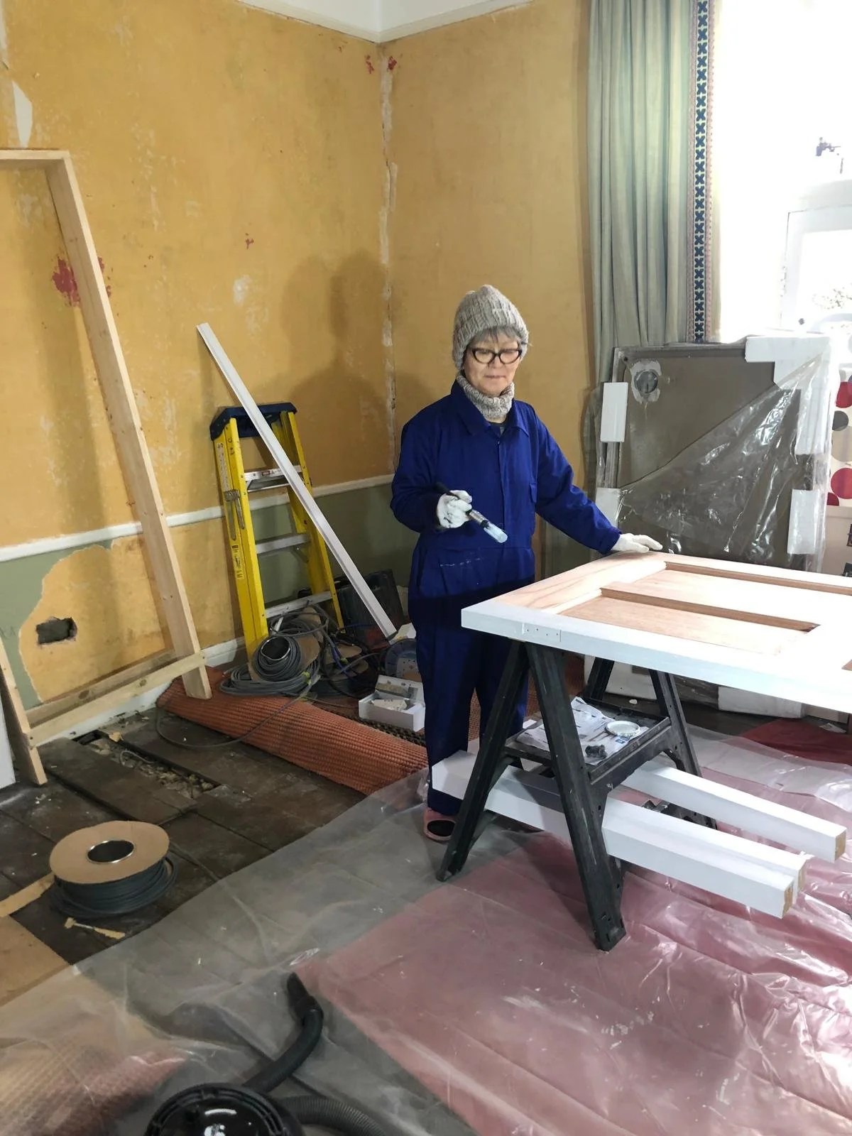 A woman wearing glasses, a gray knit hat, and a blue work jumpsuit, holding a paint roller, standing in a room under renovation with partially removed wallpaper and construction tools nearby.