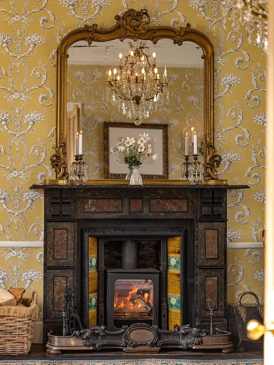A vintage living room with a black fireplace, a gold-framed mirror, a chandelier, and a floral wallpapered wall. The fireplace has a fire burning, and a vase with flowers sits on the mantel. Two candlesticks with lit candles flank the mirror.