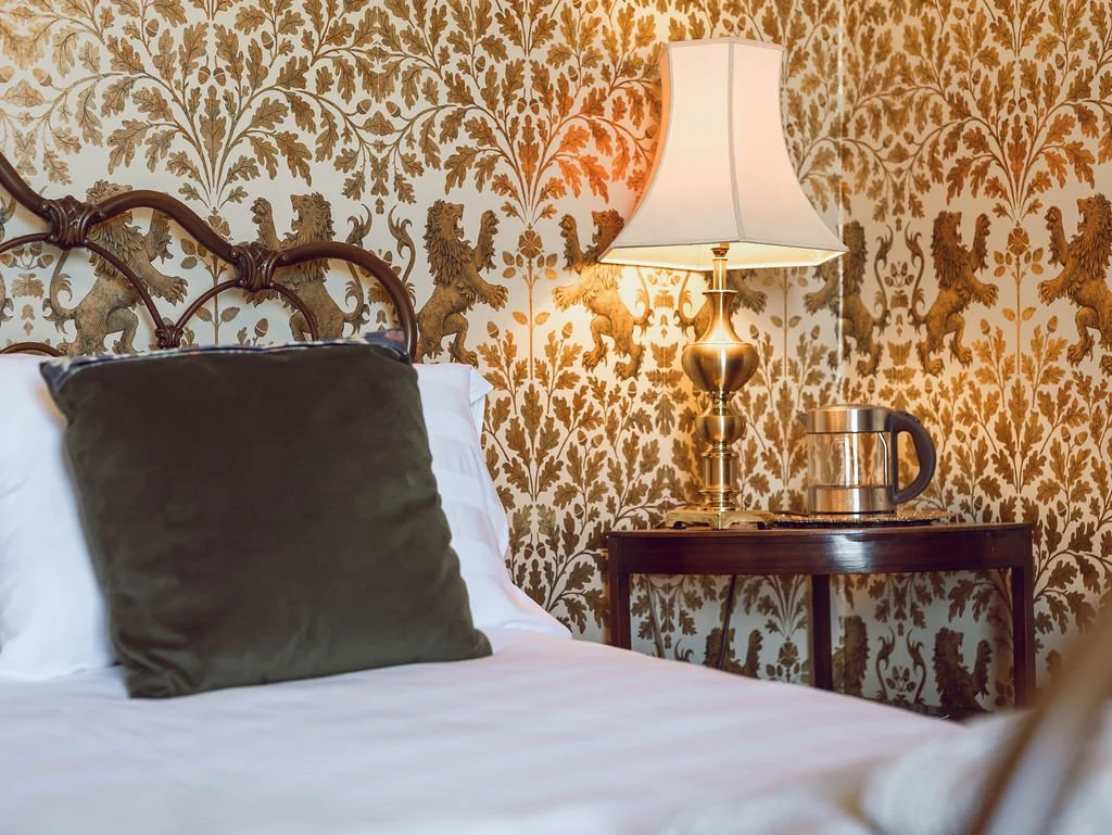A bedroom with a bed, a dark pillow, ornate wallpaper with lion motifs, a brass table lamp, and a wooden side table with a water pitcher.