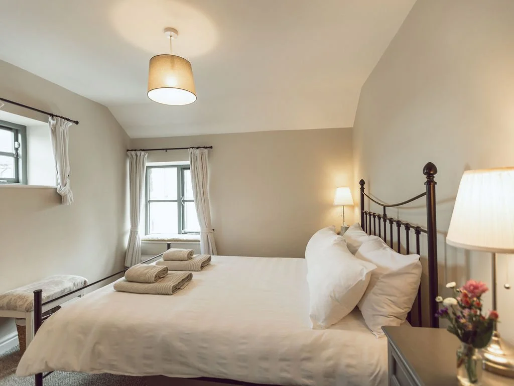 A cozy bedroom with a metal bed frame, white bedding, and pillows. Towels are neatly folded on the bed. There are two small windows with white curtains, a bedside table with lamps, and a small floral arrangement. The room has a beige ceiling and wall