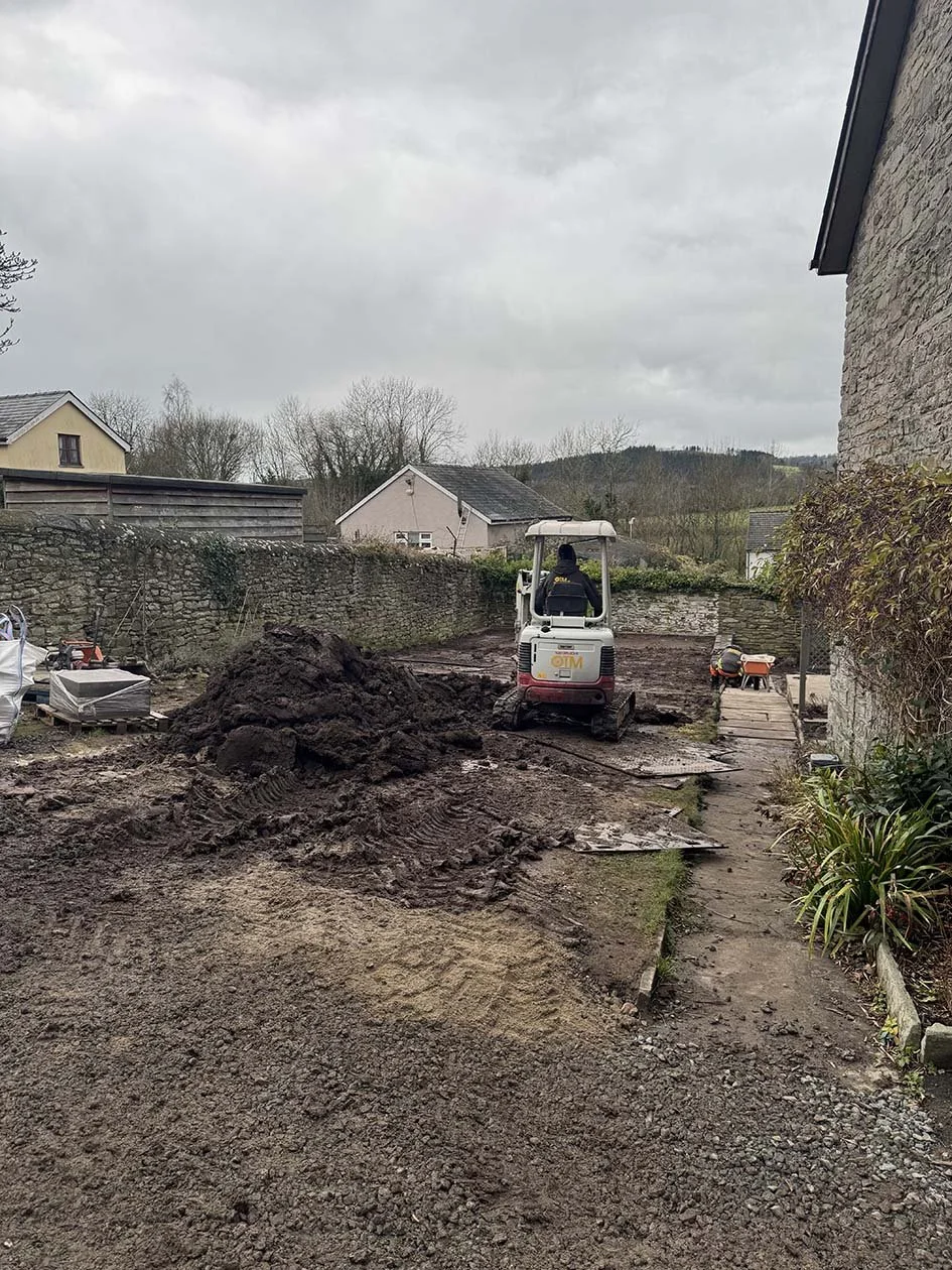 A construction site with a small excavator moving soil, a worker in the background, and a stone wall with nearby houses under an overcast sky.