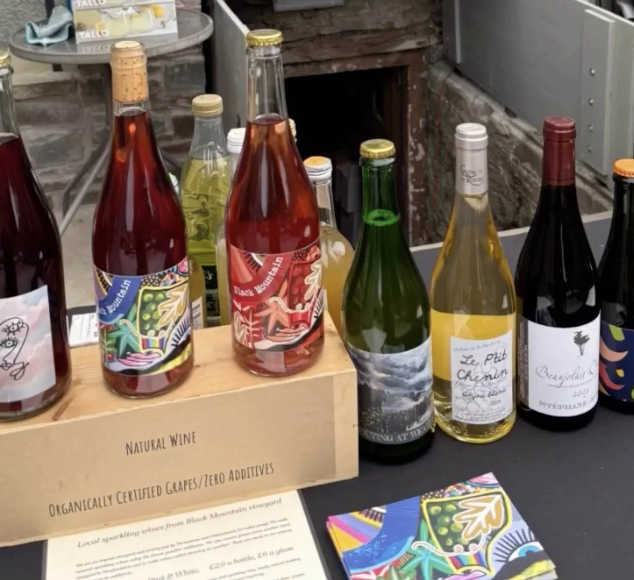 A display of colorful bottles of natural wine on a table at a tasting event, with a wooden box labeled 'Natural Wine' and 'Organically Certified Grapes / Zero Additives'.