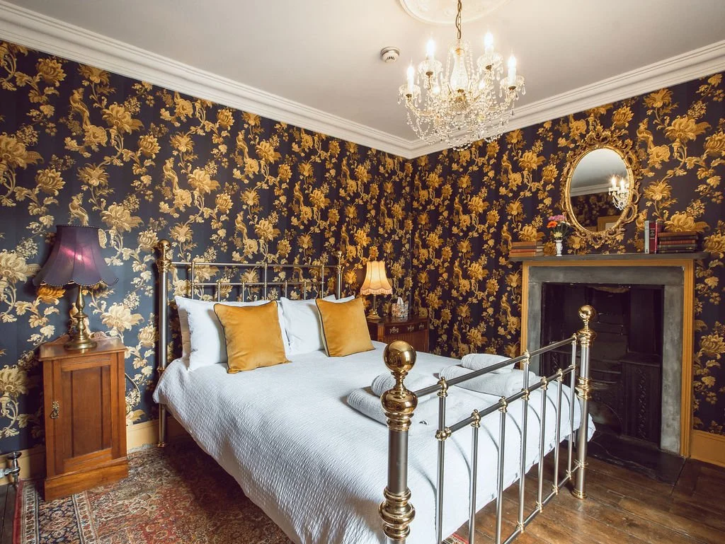 A bedroom with ornate floral wallpaper in black and gold, a brass and iron bed with white bedding and gold pillows, wooden nightstands with lamps, a fireplace with a mirror and books above it, a chandelier hanging from the ceiling, and a patterned ru