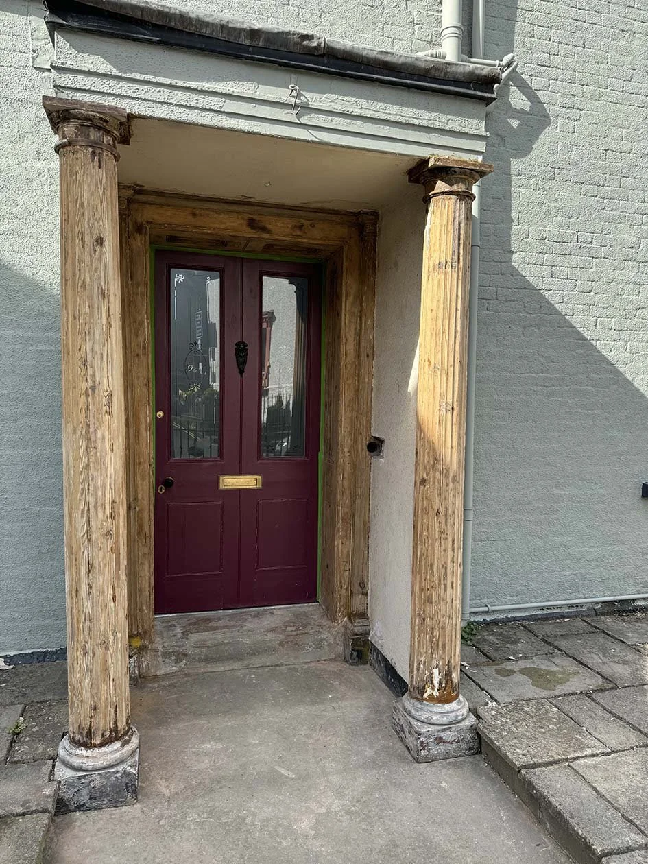 Front door with a wooden porch and two pillars, maroon door with glass panels, and brass mail slot, in a light-colored brick building.