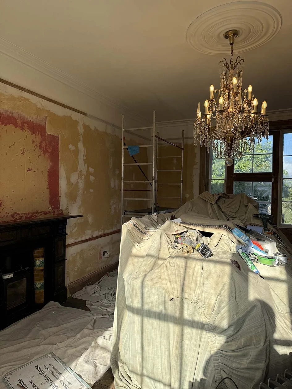 Living room undergoing renovation with wall prepared for wallpaper, cluttered furniture, and a chandelier hanging from the ceiling.