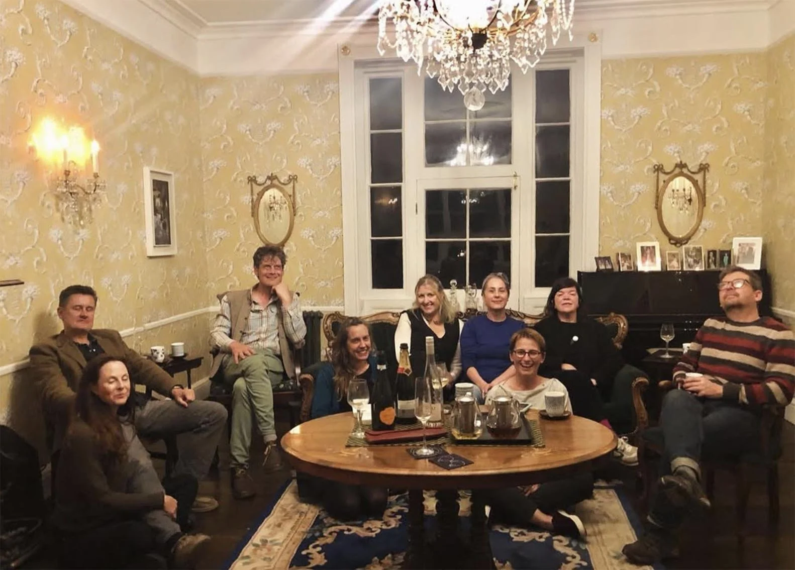 A group of nine people gathered in a well-decorated living room, sitting and standing around a coffee table with drinks and bottles. The room features vintage wallpaper, a chandelier, framed photos, and a grand piano.