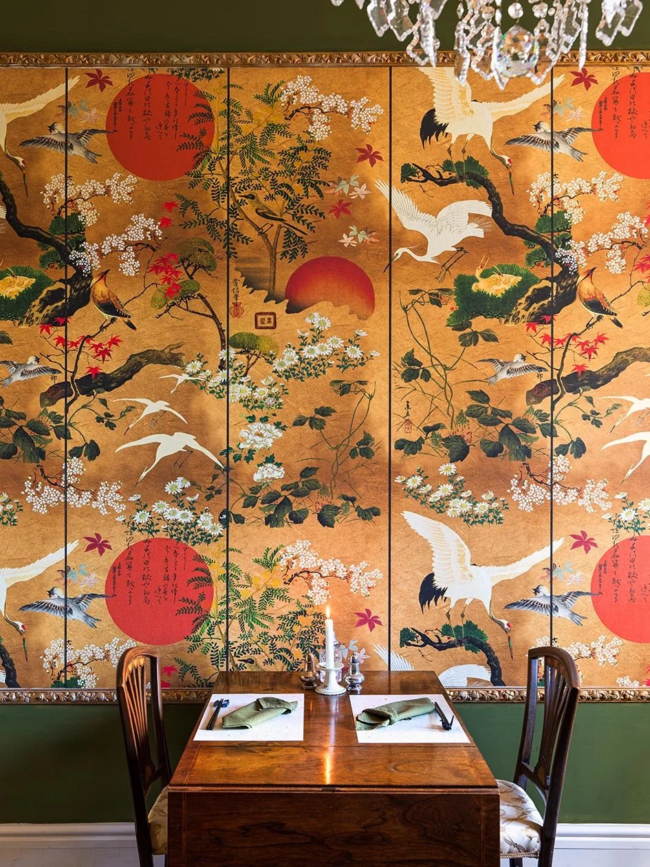 A dining table set for two with folded green napkins, utensils, a lit candle, and salt and pepper shakers, in front of a decorative Japanese-style screen featuring cranes, cherry blossoms, and red sun motifs.