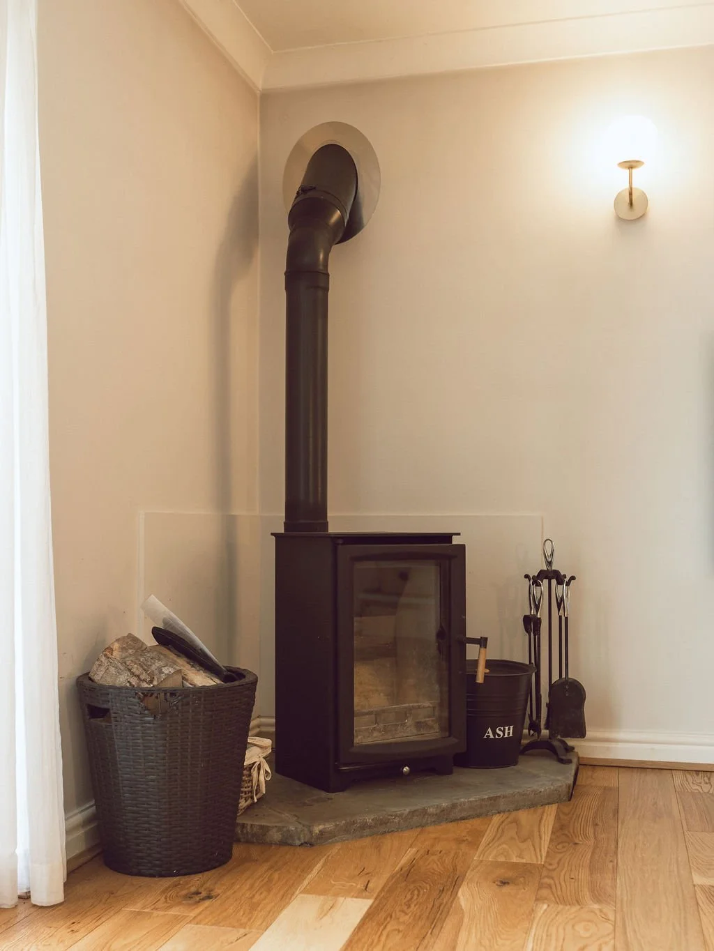 A black wood stove with a glass door sits on a stone hearth in a room with light-colored walls. To the left is a basket filled with firewood, and to the right are fireplace tools and a bucket labeled 'ASH'. A wall sconce provides warm lighting in the