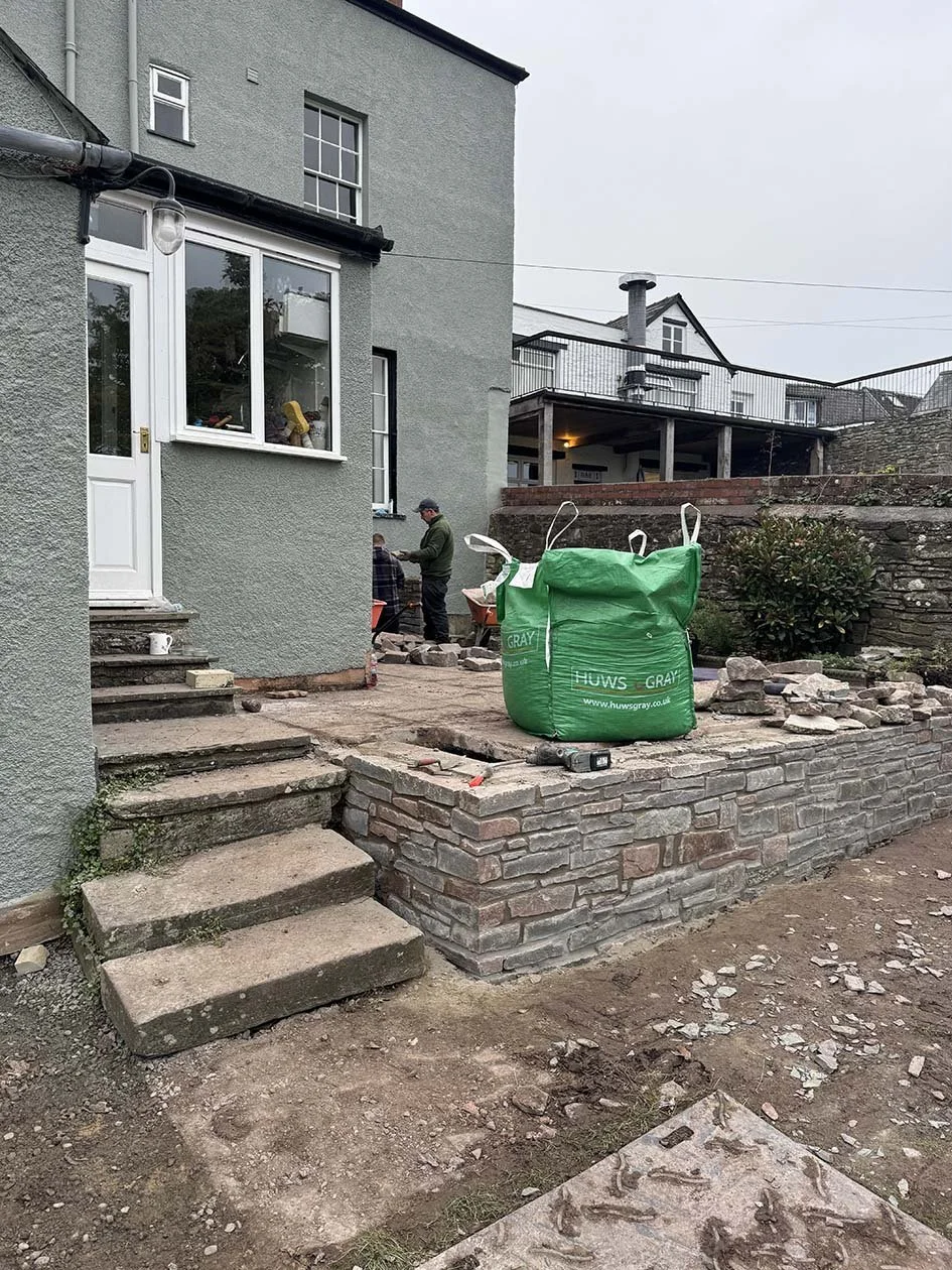 Construction workers are building a new outdoor patio or steps area at a house, with bricks and paving stones, and green bags of construction materials nearby.