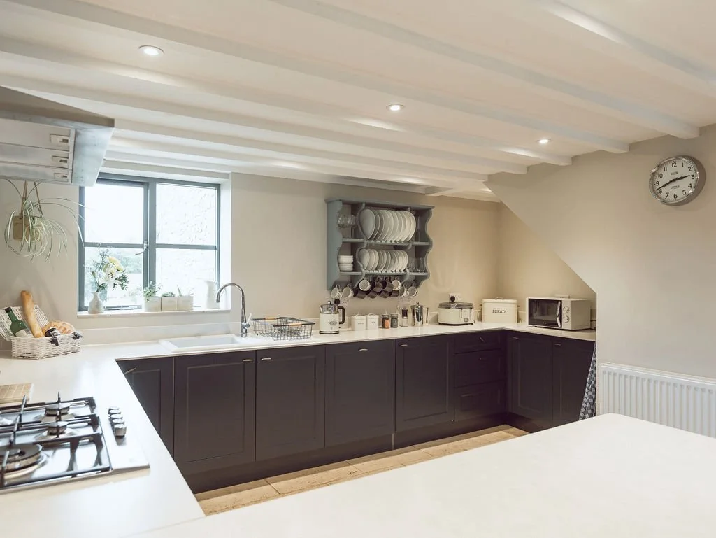 A modern kitchen with white countertops, dark cabinets, and a window above the sink. The kitchen has open shelves with plates, a microwave, a bread box, and various small appliances. There is a clock on the wall and a radiator.