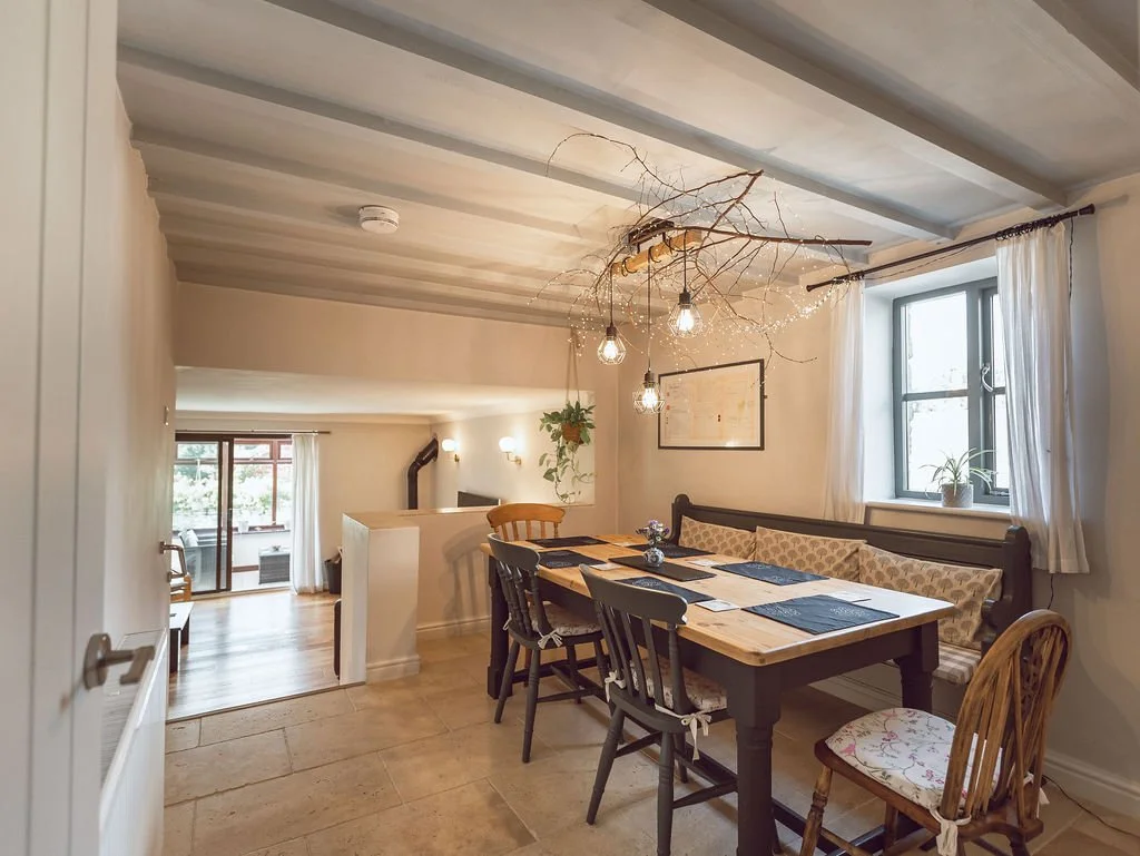 A dining area with a wooden table, various chairs, a built-in bench with patterned cushions, and a window with white curtains. Above the table hangs a branch with exposed light bulbs and fairy lights, creating a cozy atmosphere.