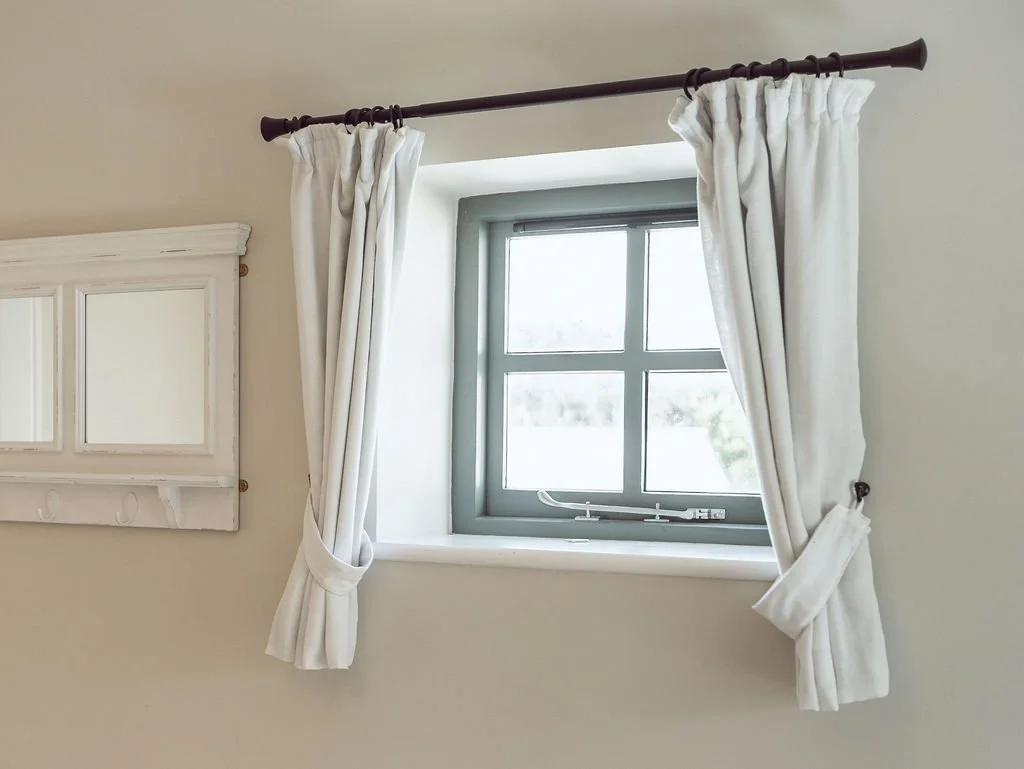 A small window with four panes, framed in gray, with white curtains held back with tiebacks, and a white wall with a white wall-mounted cabinet nearby.