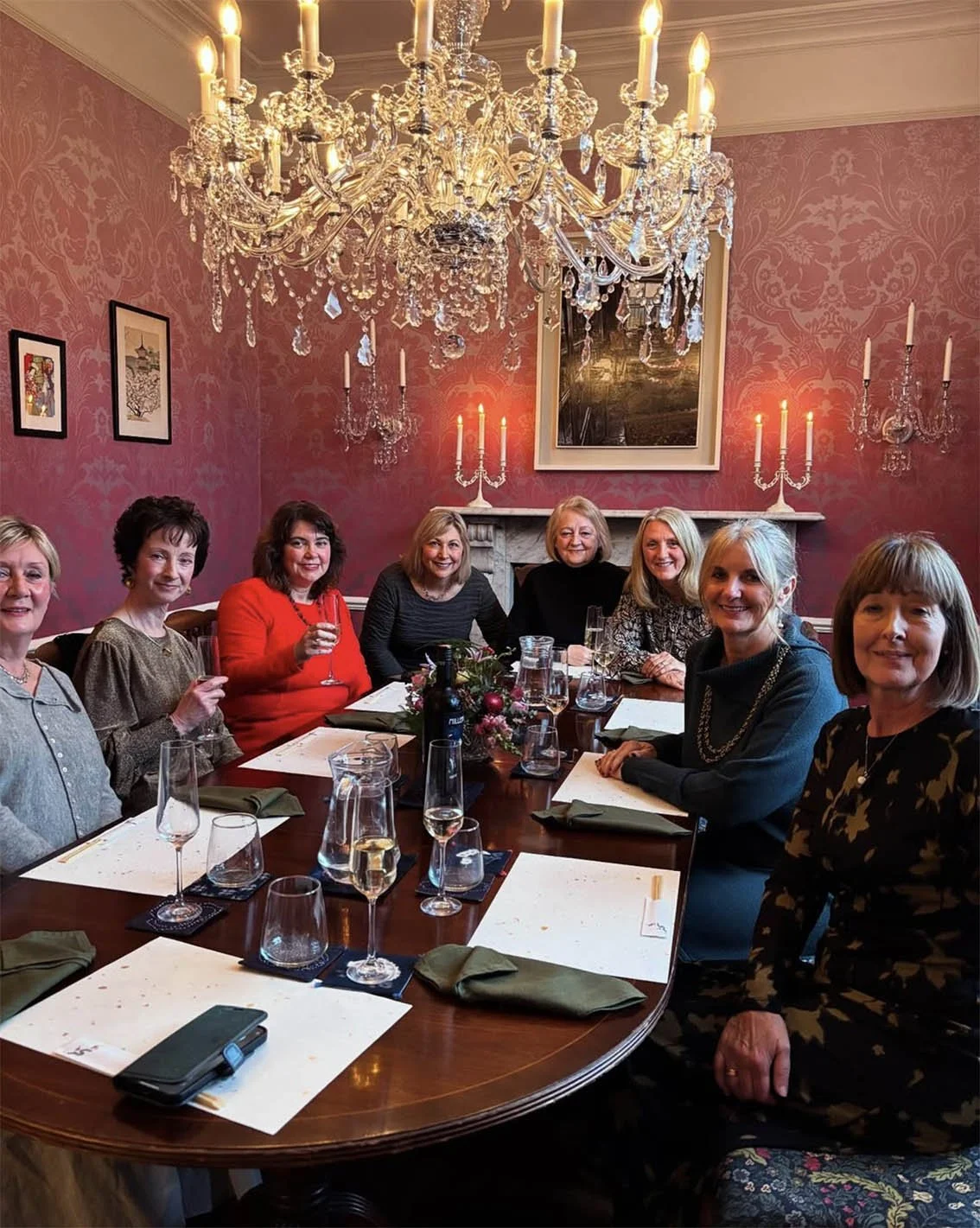 A group of women sitting around a dining table in an elegant room with pink wallpaper, a large chandelier, and candle wall sconces.