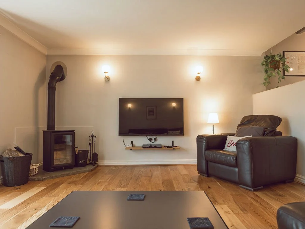 Living room with a wood-burning stove, a wall-mounted TV, a leather armchair with a pillow, a lamp, and a small table.
