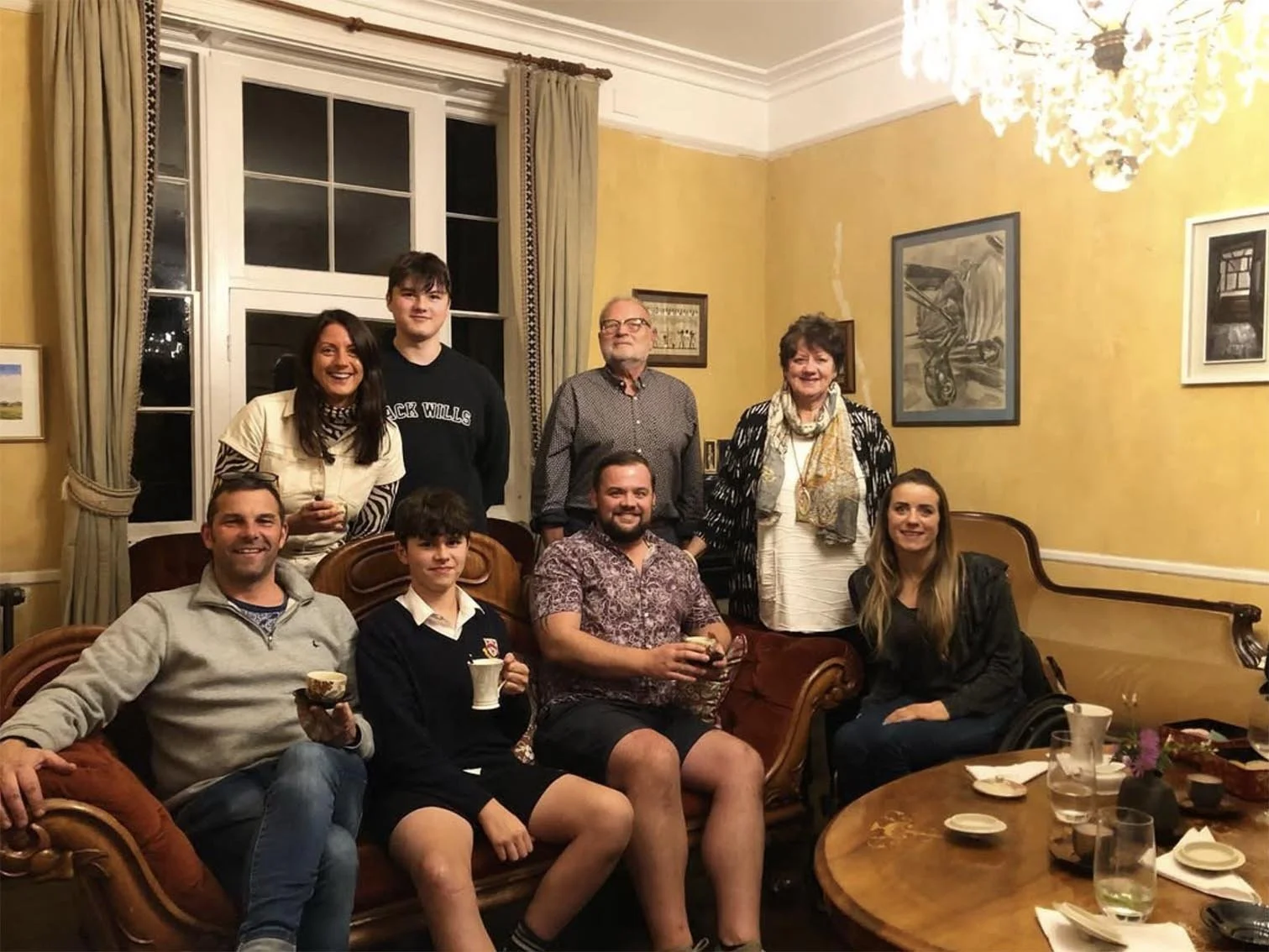 A group of nine people gathered in a warmly decorated dining room, smiling at the camera. Some are seated on a sofa around a table with cups and plates, while others stand behind them. The room has yellow walls with framed artwork, large window with 