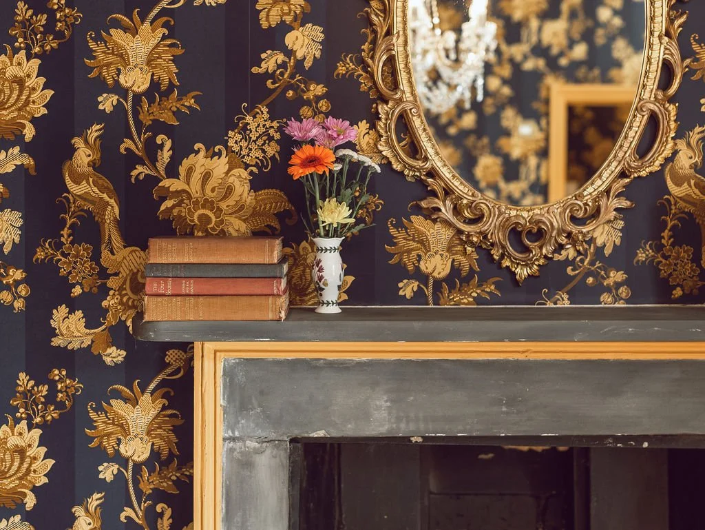 A decorative shelf above a fireplace with a vase of flowers and stacked antique books, a large ornate gold mirror on dark floral wallpaper.