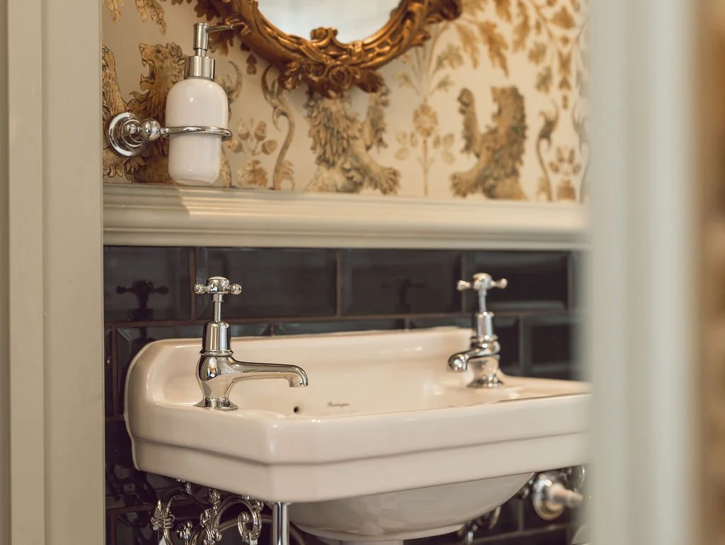 Close-up of a vintage white porcelain double sink with chrome faucets in a bathroom, above black tiled wall with ornate wallpaper and mirror.