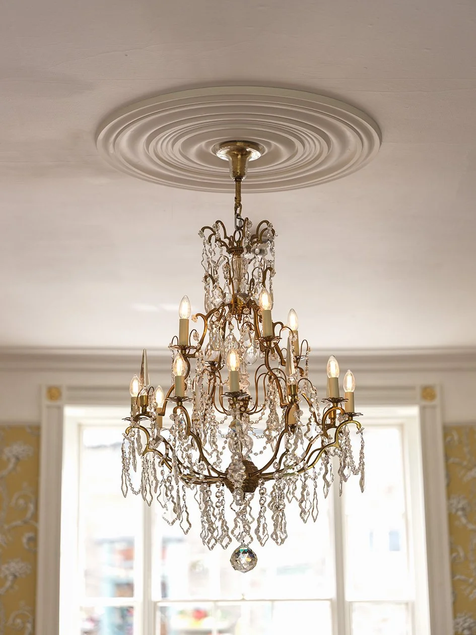 A gold and crystal chandelier hanging from an ornate ceiling medallion in a bright room with large windows.
