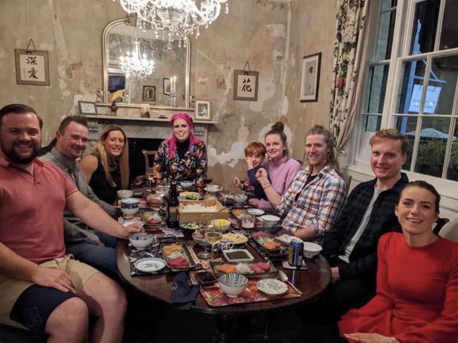 Group of people sitting at a long dinner table with food, drinks, and chopsticks in a cozy, decorated room