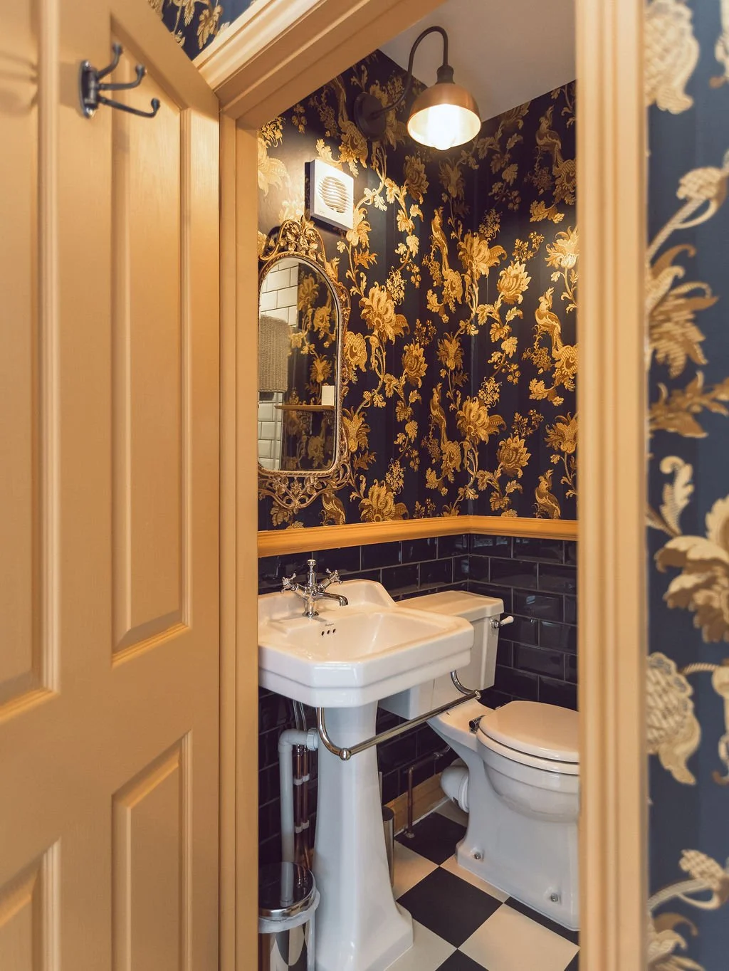 View of a small vintage-style bathroom with black and gold floral wallpaper, a white pedestal sink, a toilet, a gold-framed mirror, and a black and white checkered floor, seen through an opened door.