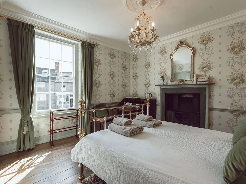 A bedroom with a large window, green curtains, a chandelier, a bed with rolled towels, a fireplace with a mirror above it, and vintage wallpaper.