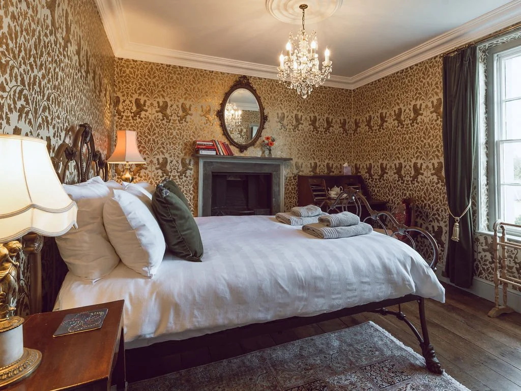 A vintage-style bedroom with a large bed, ornate headboard, and white bedding, accented by several pillows. There are open towels on the bed. The room has intricate gold and brown floral wallpaper, a chandelier hanging from the ceiling, and a window 