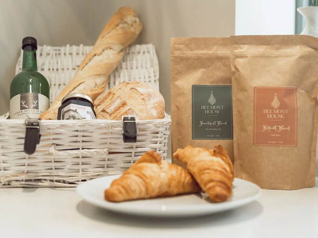 Bread, croissants, a bottle of cider apple juice, bags of Belmont House breakfast blend coffee, and a plate with croissants on a white table.