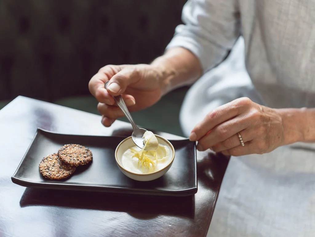 Person serving cream or butter from a small bowl onto a plate with two cookies.