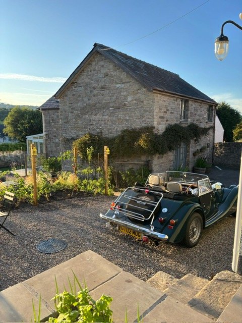 A vintage convertible car parked outside a stone house with a small garden, in a rural setting during late afternoon or early evening.