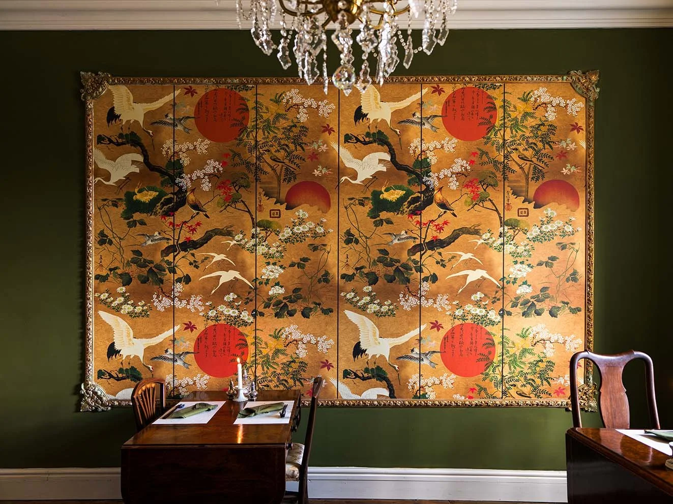 Decorative Asian-style folding screen with cranes, red sun, and floral patterns hanging on a dark green wall in a dining room with wooden tables and chairs, topped by a crystal chandelier.