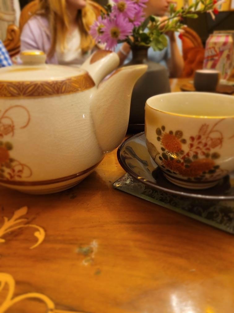 Teapot and teacup on a wooden table, with a vase of pink flowers and blurred people in the background.