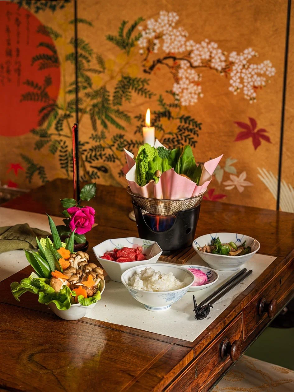 A traditional Japanese table setting featuring a hotpot with vegetables, flowers, rice, and side dishes on a wooden table with a decorative screen in the background.