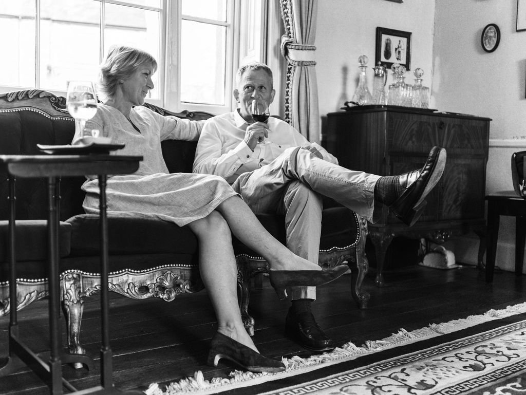 A black and white photo of an older woman and a man sitting on a vintage sofa. The woman appears to be talking to the man, who is drinking from a wine glass, in a cozy living room with framed pictures on the wall and a window with curtains.