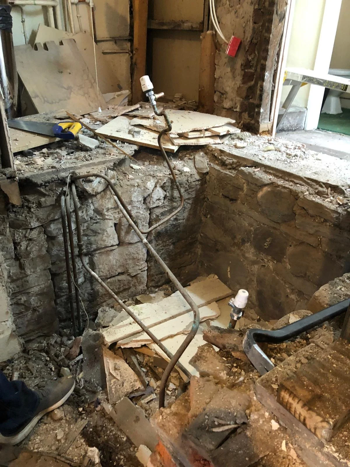 A construction site showing an open wall with exposed bricks and plumbing pipes, surrounded by debris and tools, indicating ongoing renovation work.