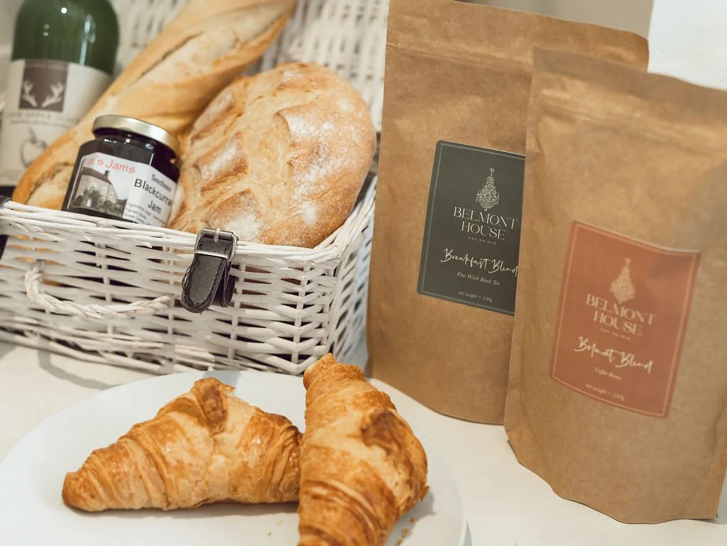 A breakfast spread with two croissants on a white plate, a basket of assorted bread including baguette and rolls, a jar of blackberry jam, and two brown paper bags labeled 'Belmont House Breakfast Blend' coffee and tea.