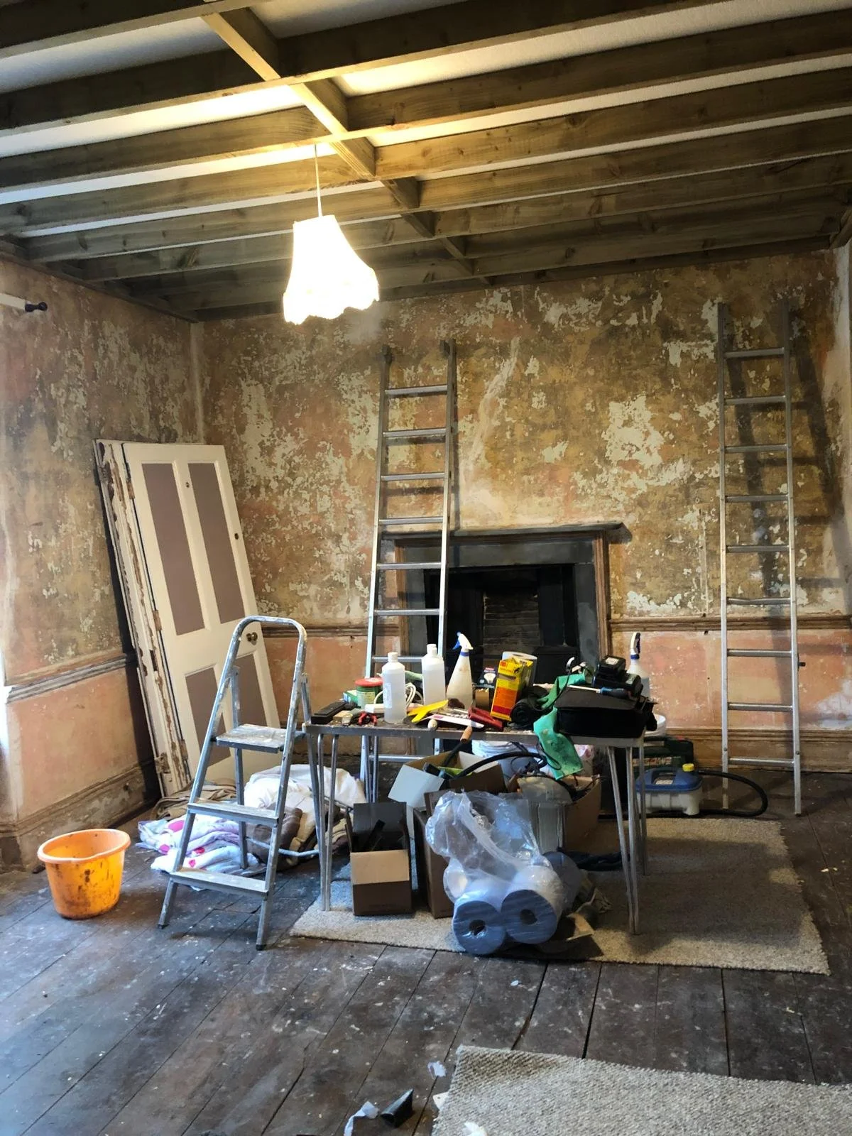 A room under renovation with walls stripped and peeling paint, wooden floors, two aluminum ladders leaning against the wall, a table cluttered with tools and supplies, a door resting against the wall, and a partially visible fire place.