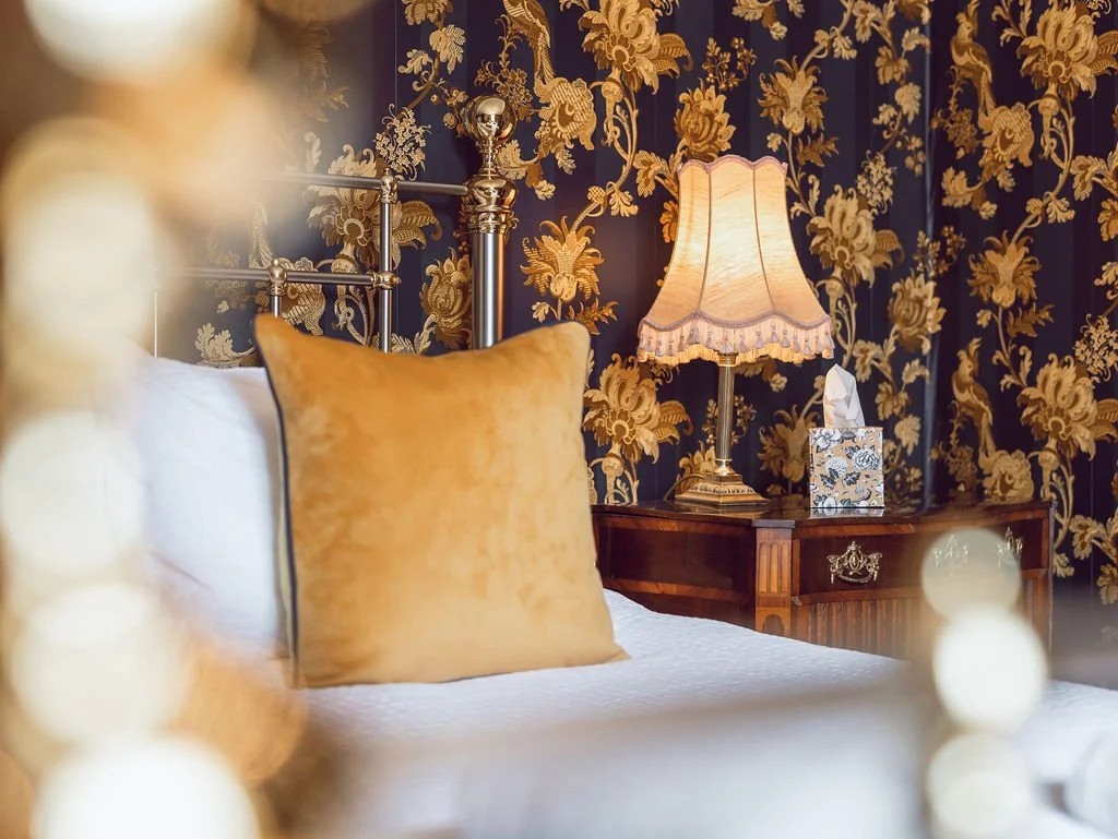 A luxurious bedroom with a bed that has white sheets and a golden pillow, a vintage wooden nightstand with an ornate drawer handle, a decorative table lamp with a fabric shade, and floral wallpaper with gold and dark blue patterns.